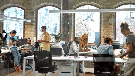 Several people are sitting in an office behind a glass wall working and partly chatting